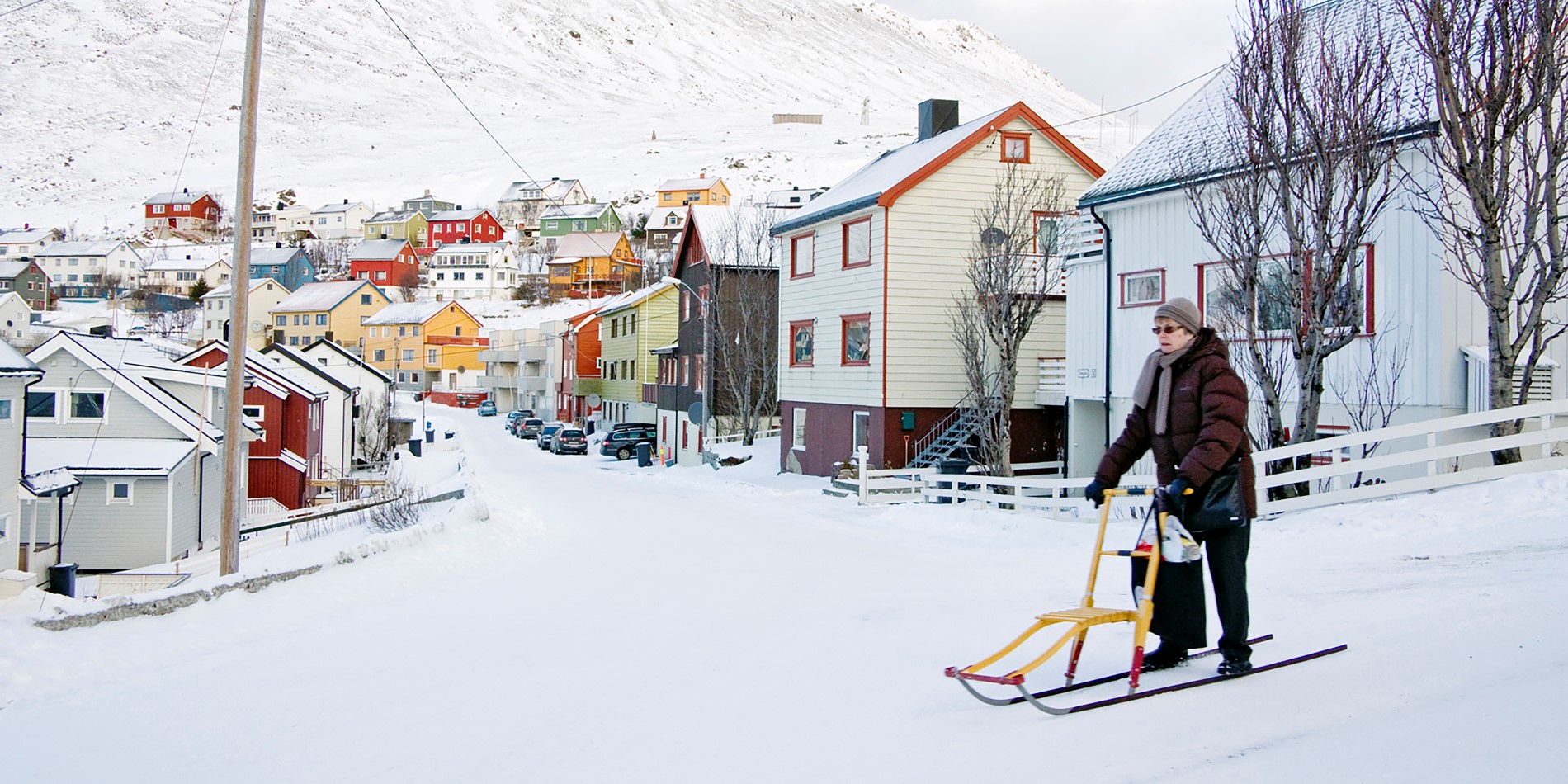 Billedet viser en ældre kvinde i sneen foran en gade med smukke og farverige, små træhuse