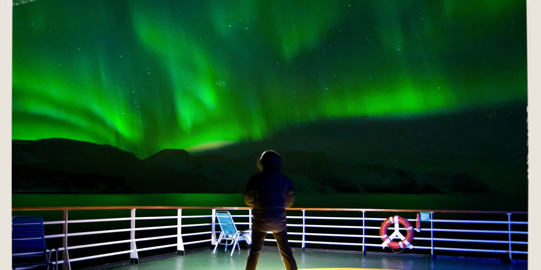 Mand står på dækket af Hurtigrutens Den norske kystrute. Over ham er himlen oplyst i grøn – nordlyset danser