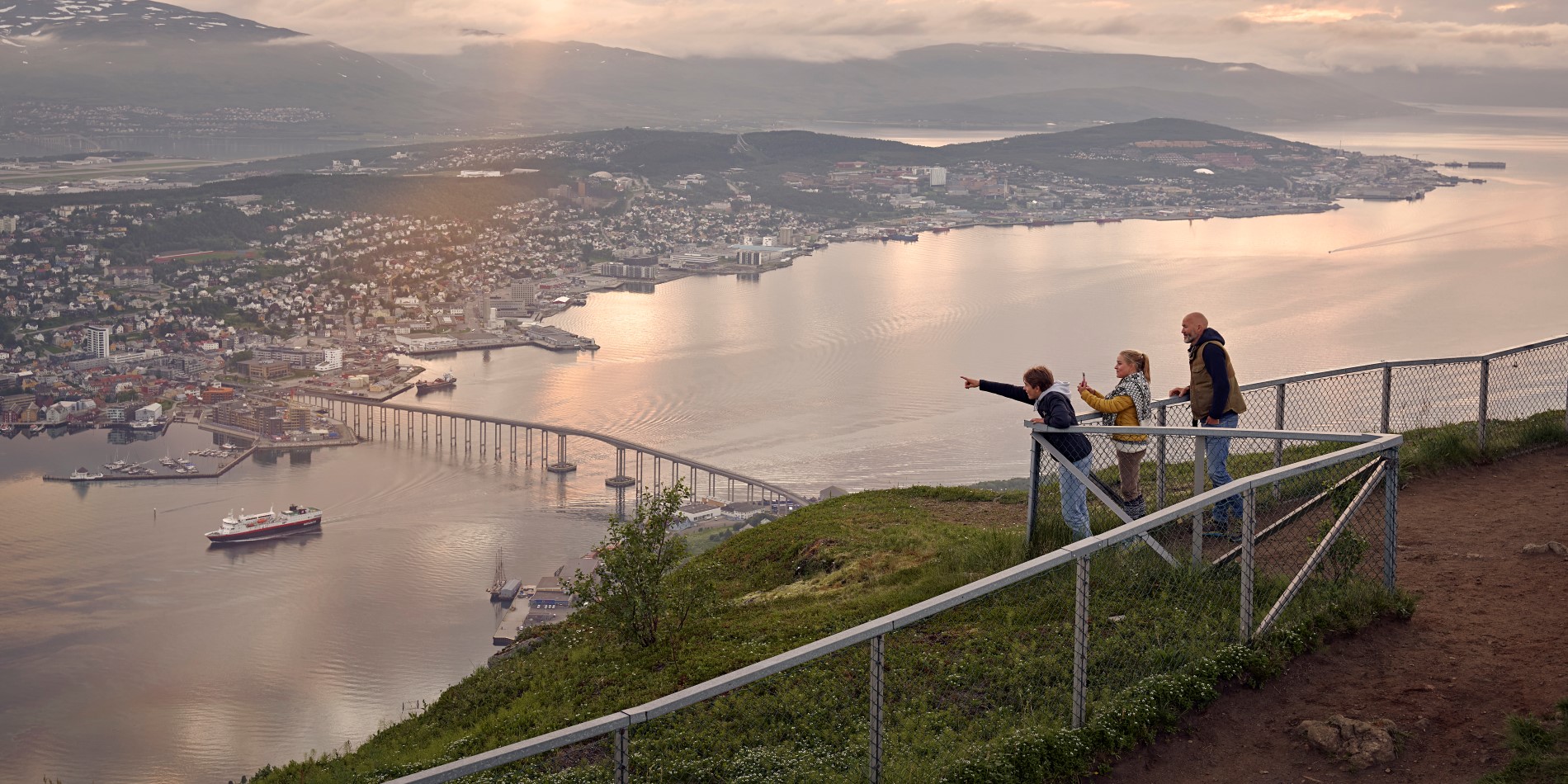 Tre børn står bag et hegn på toppen af Tromsøs byfjeld. De ser ud over Tromsø by.