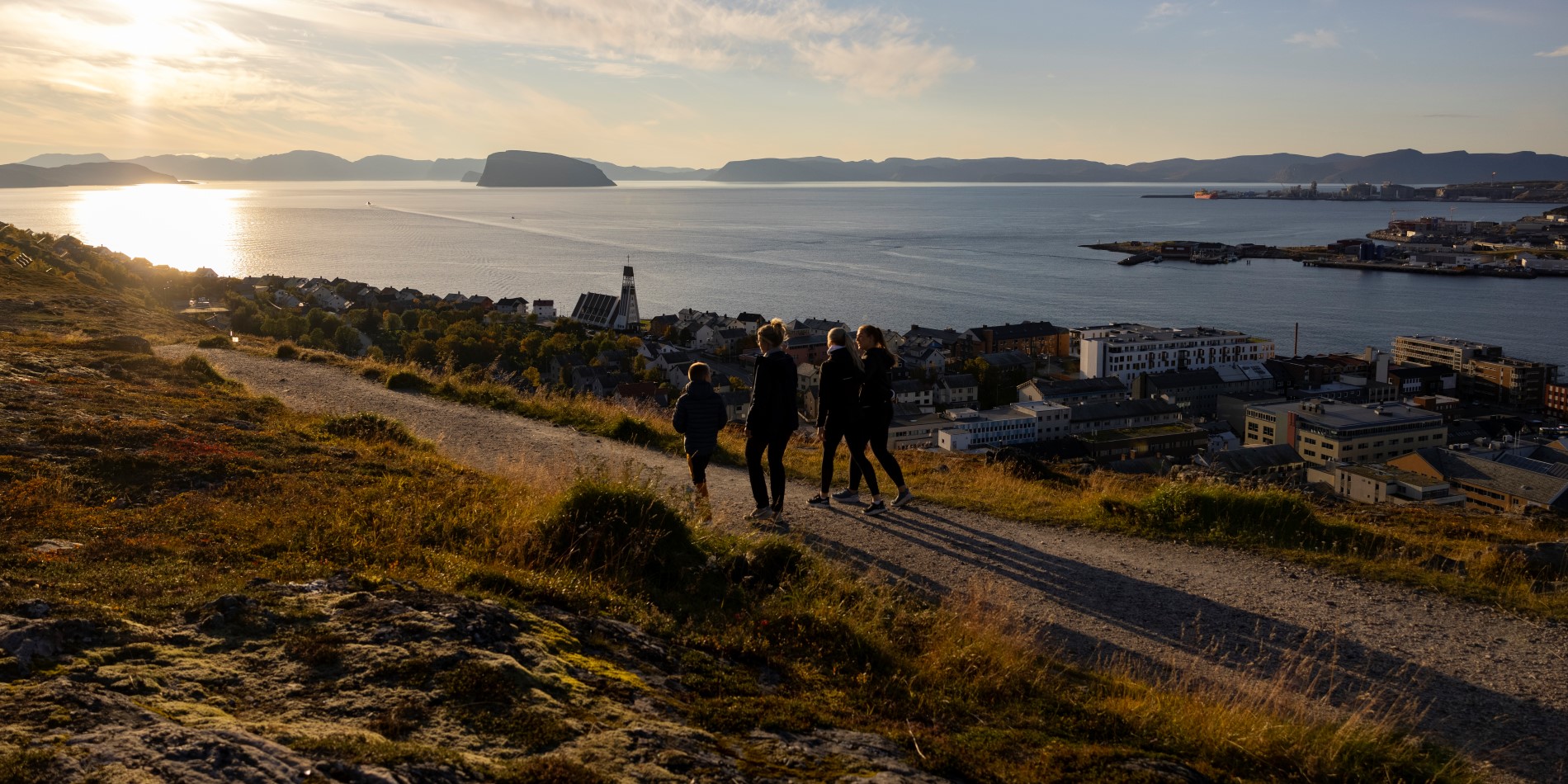 Det er solnedgang, og fire mennesker går på fjeldsiden. Under dem kan man se Hammerfest
